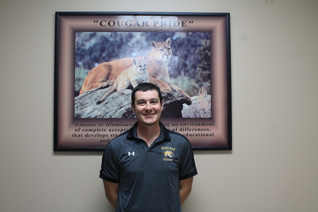 A smiling Michael Scoggin, wearing a dark grey Under Armour polo shirt with a "HIGHLAND COUGARS" logo,  stands in front of a framed "COUGAR PRIDE" poster featuring a photo of a cougar and cub.
