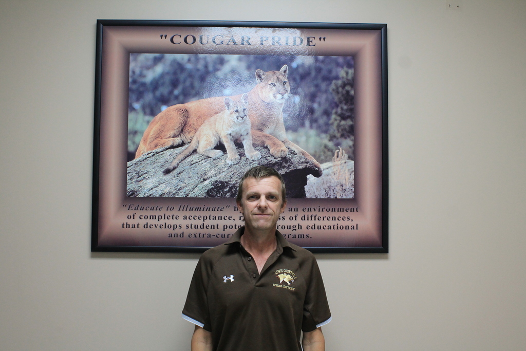 A picture of John Sparks, wearing a brown polo shirt with Under Armour and school district logo, standing in front of a large framed "COUGAR PRIDE" poster featuring a cougar family and educational text.