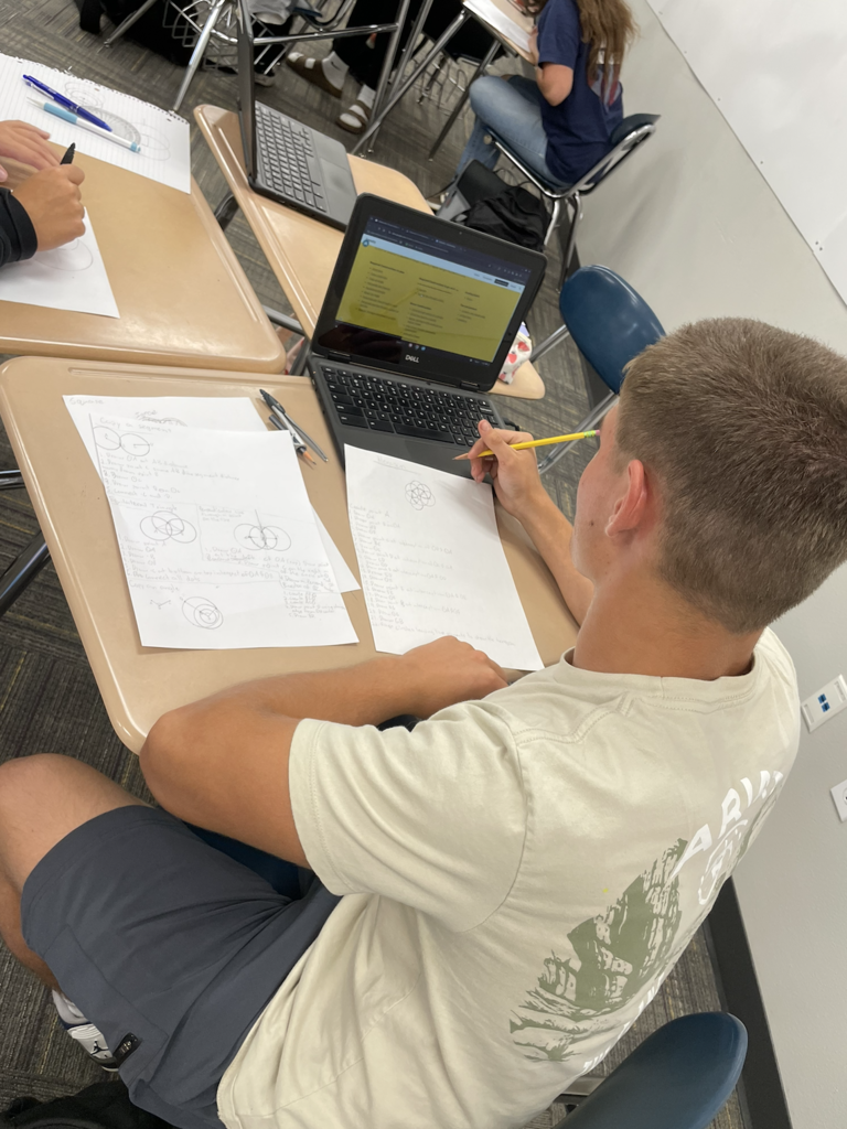 student working on a paper and a computer screen, student is wearing a beige shirt and grey shorts