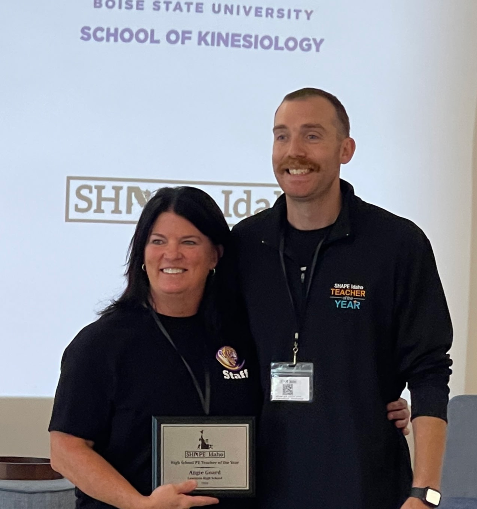 woman with brown hair and black shirt receiving a plaque from a man with a black sweatshirt and black shirt on with a mustache and short brown hair