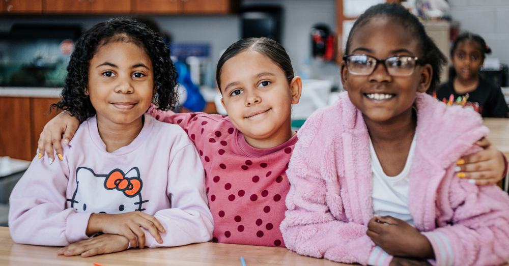 Three smiling Charles England Elementary School students sit together at a classroom table, with their arms around one another.