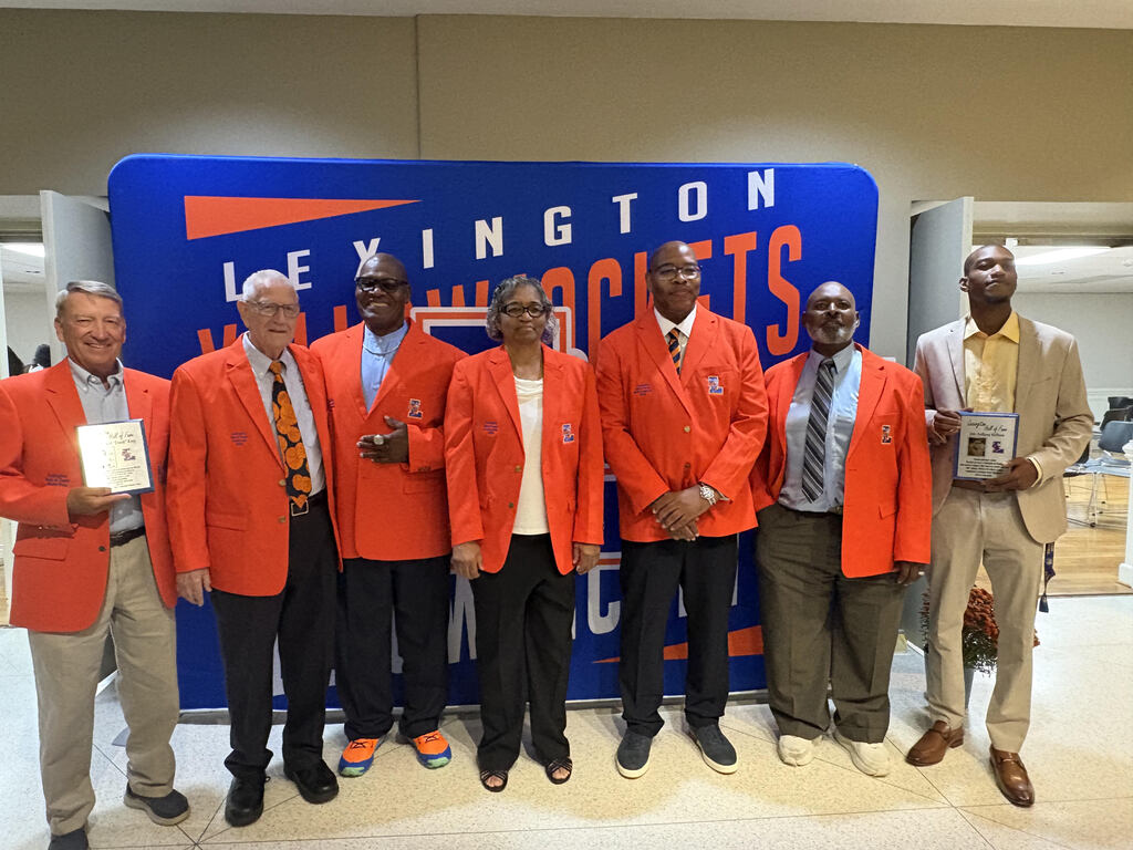 Lexington Senior High School Athletic Hall of Fame Class of 2026 inductees pose for a group photograph during their induction ceremony, with several wearing orange jackets and holding commemorative plaques in front of a Lexington Yellow Jackets backdrop.