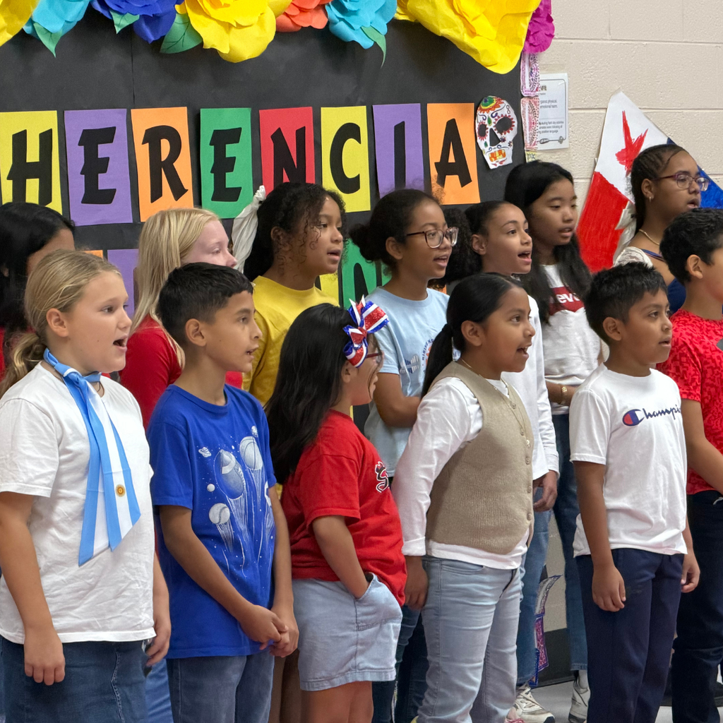 Pickett Elementary School students sing together in front of a colorful Hispanic Heritage Month display featuring flags and cultural decorations.