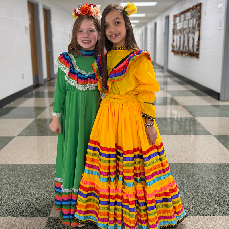 Two Pickett Elementary School students smile while wearing colorful traditional dresses during the school's Hispanic Heritage Month celebration.