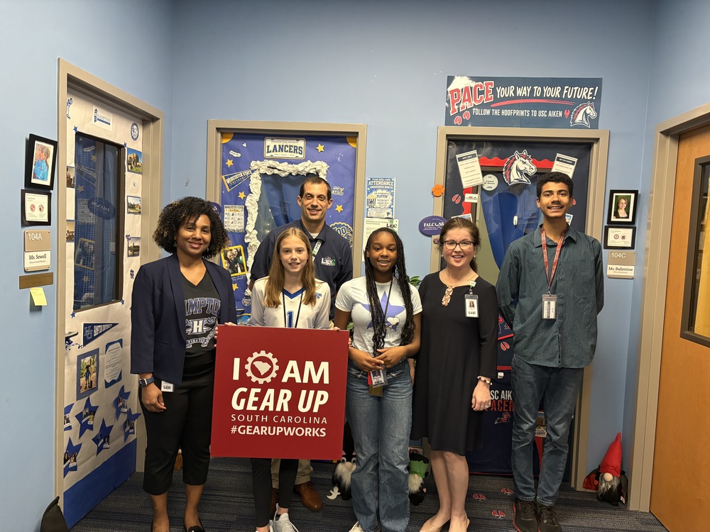 A group of students and adults who assessed and voted on different doors decorated in college information are standing together smiling at the camera. One student in the front of the group photo is holding a square and red sign that states, "I am Gear Up, South Carolina #GearUpWorks.