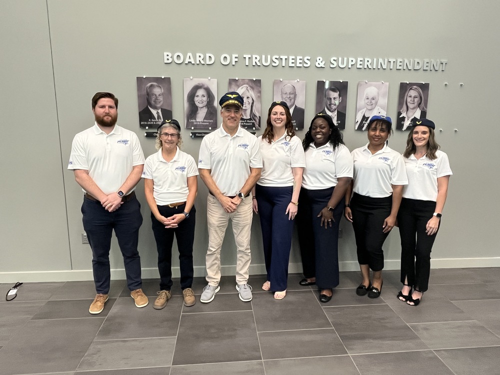 A group photo of FMS's Administration team, School Counselors, Instructional Coach, and School Social Worker. They are standing together facing and smiling at the camera. They are standing in front of photos on the wall of the Board of Trustees and Superintendent. They are dressed in white collared three button shirts that state "Flight" on them. Some are wearing hats that pilots and airplane staff members wear.