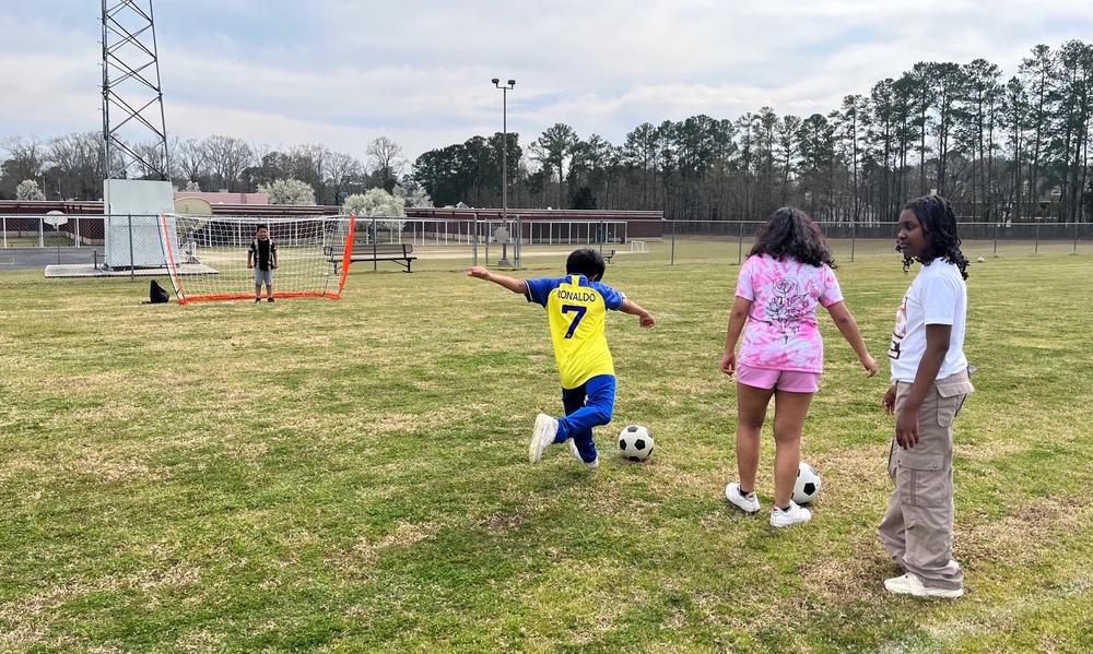 Photo of 3 students facing soccer goal, one student kicking ball toward goal