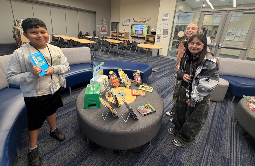 Three students are shown standing next to the "Butter Book Display" table in the library next to the couches. This display has books on and off of easels. There are slices over butter on "book peepers" peeking out of some of the books. The display sign about "Spread the Love of Reading" and "You Better Believe these are google books" are in clear acrylic stands sitting on top of upside down storage bins so students can view the signage. The students are smiling at the camera.