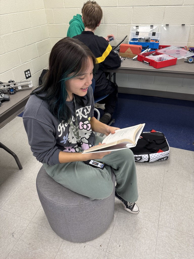 A female student sitting on an ottoman during class and reading a book.