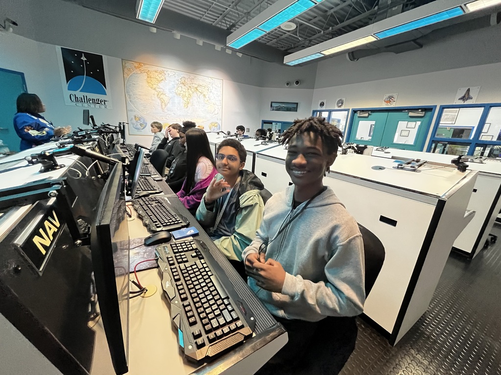 Students smile for the camera while sitting at a computer table inside of a challenger space control center.