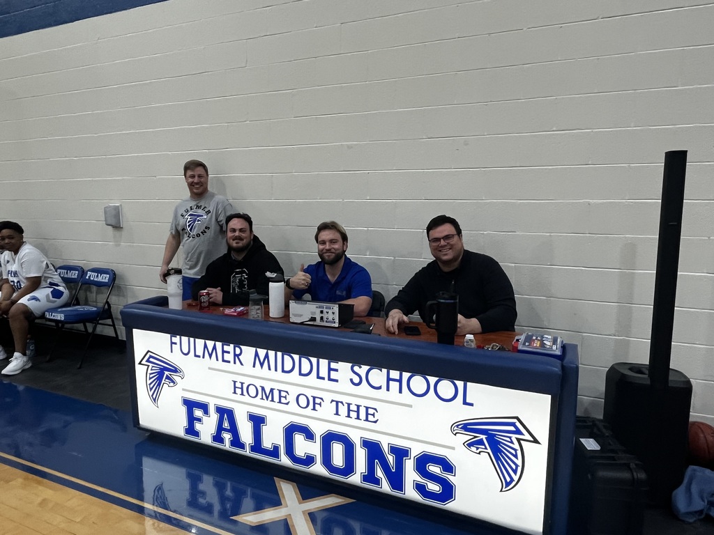 Three staff members sit and one stands at the press box during half time of the basketball game. They are smiling for the camera.