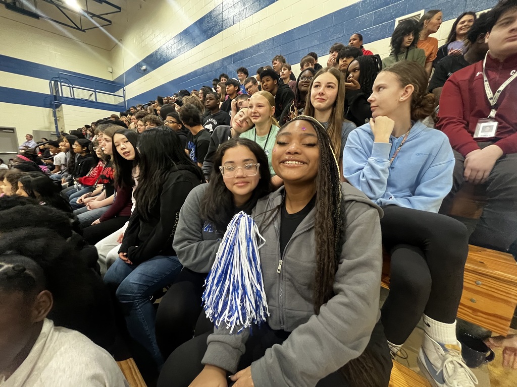 Two girls smile for the camera while sitting in the bleachers. Other students are in the background sitting in the bleachers.