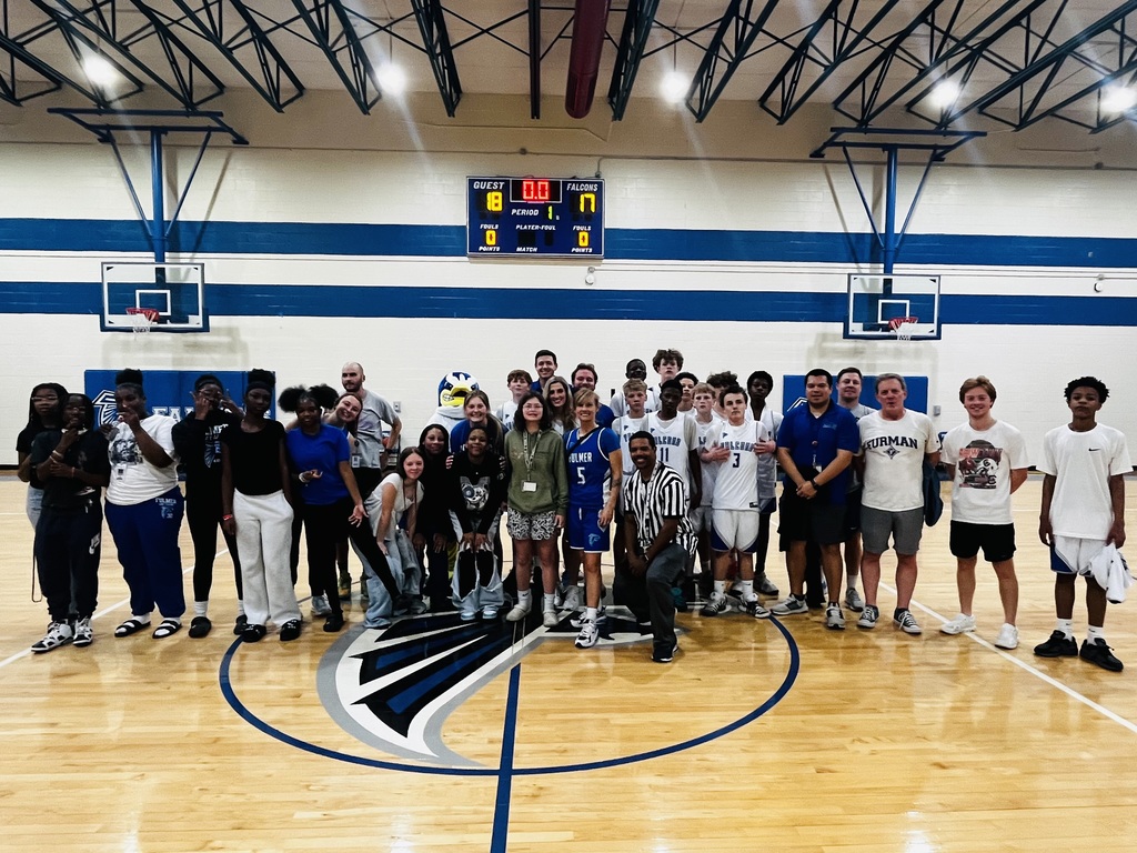All team members to include faculty/staff and students who participated in the faculty/staff vs. student basketball game stand in the middle of the gym and smile for the camera taking a group picture with the scoreboard in the background.