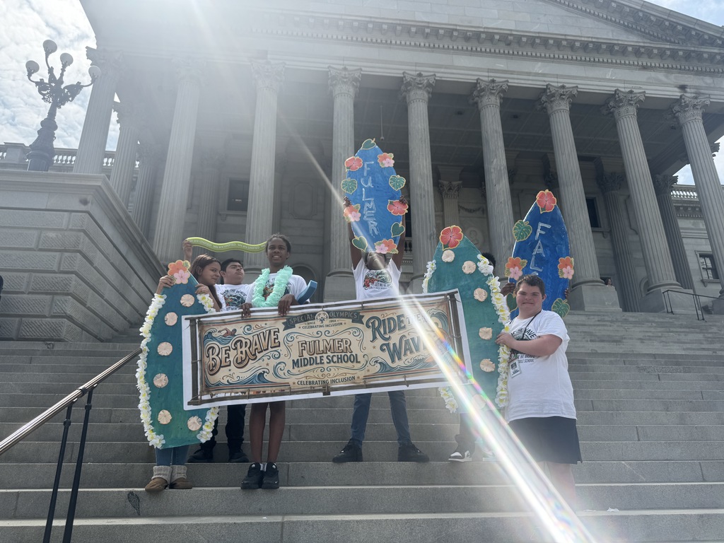 A small gruop of students hold a banner that states "Be Brave, Ride the Wave , Fulmer Middle School along with holding surfboards that state Fulmer and Falcons. The students are standing on the steps of the SC State House for this picture.