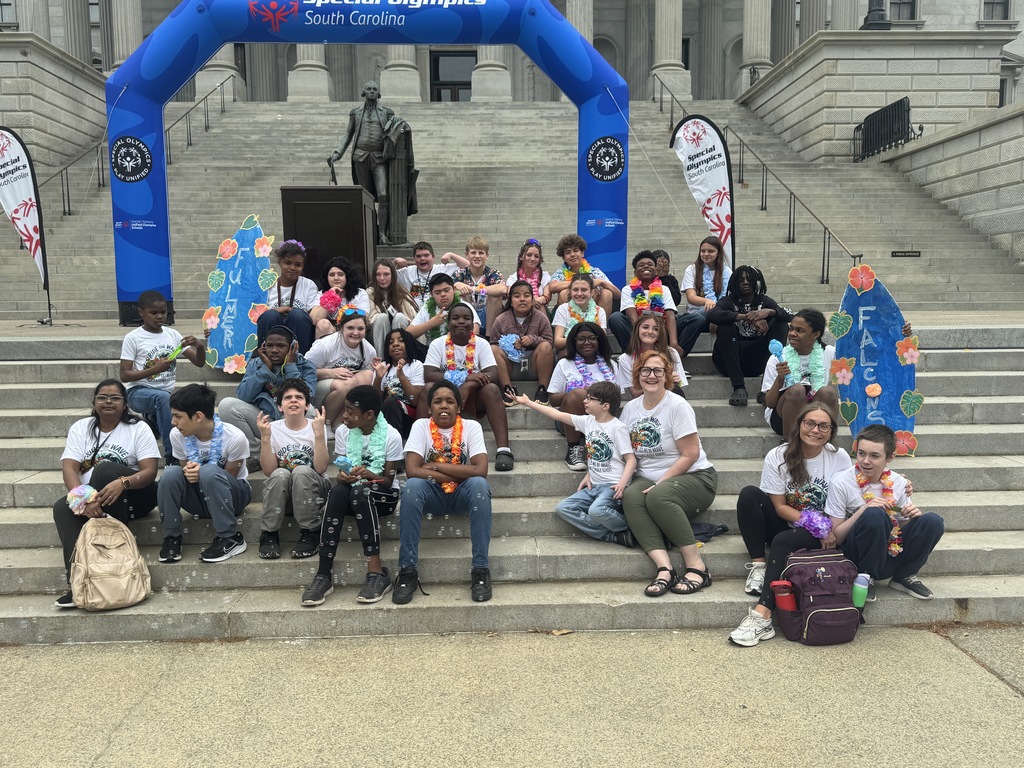 A group of students, teachers, and paraprofessionals sit on the steps of the SC State House smiling for the camera in front of the South  Carolina  Unified Rally for Special Olympics backdrop.