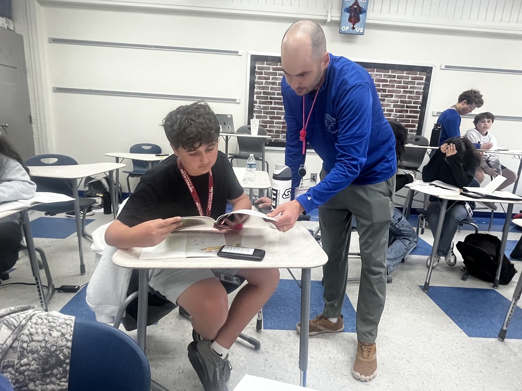 Mr. Stewart leans towards a student desk and holds the corner of a workbook the student sitting in the desk is holding as they look at a math problem together in the classroom.