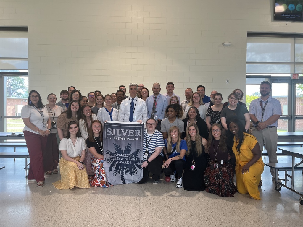 A group of R.H. Fulmer Middle School educators in the school cafeteria smile for the camera holding the Silver High Performance banner of the Palmetto Gold and Silver Award.