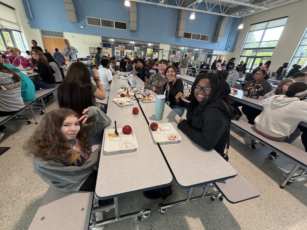 A group of students, some looking and smiling at the camera and some not, sit at the cafeteria tables during lunch.