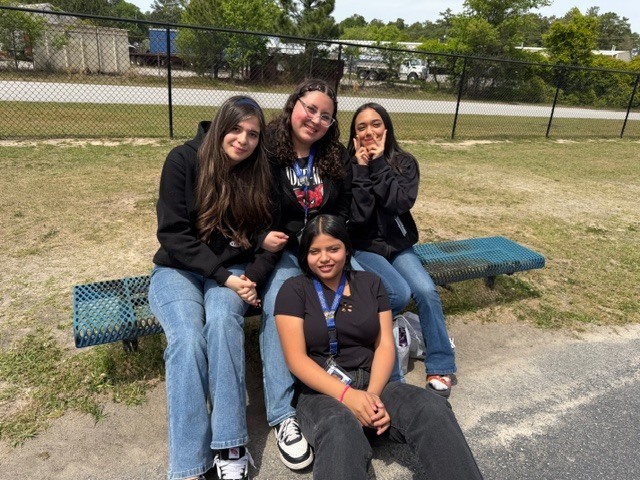 Three students sit on a bench and one on the ground outside and smile for the camera.