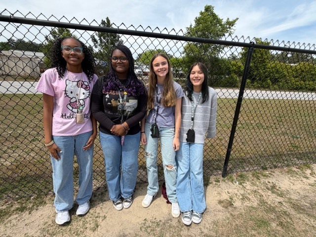 Four girls smile and stand together and lean against the fence outside during recess.