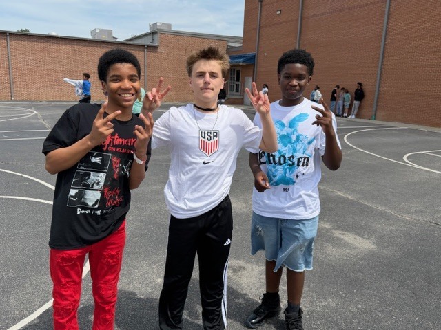 Three boys smile for the camera and hold up peace signs on the blacktop outside during recess.