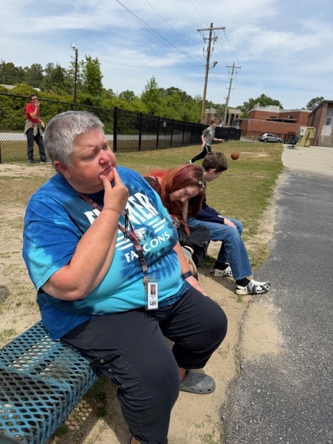 A teacher is shown sitting on a bench outside looking off into the distance with your fingers on her chin like she's watching and thinking.