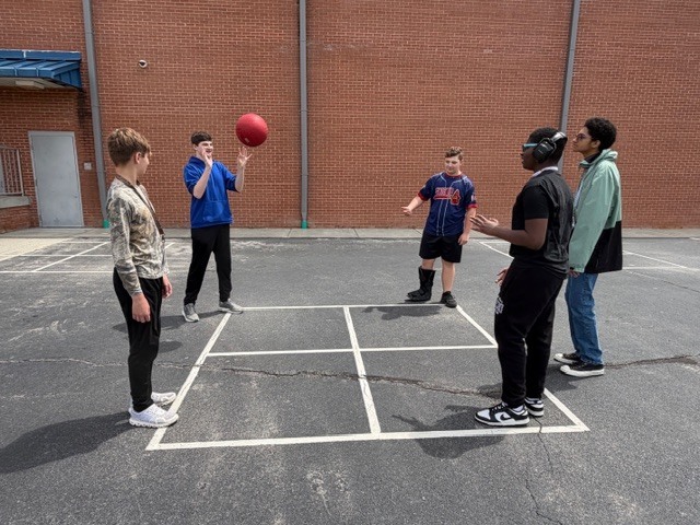 Four boys play four square while one is waiting his turn to play all on the outside blacktop during recess.