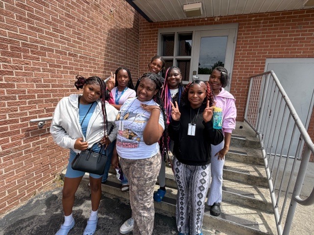A group of girls stand on the stairs and in front of the stairs outside during recess. All smile for the camera.