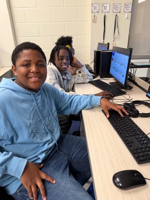 Two boy s sit at their computers in a computer lab classroom. Both smile at the camera. One student sits at his computer with headphones on looking at his screen in the background.