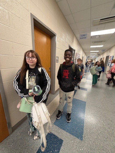 A boy and a girl stand in the hallway and smile for the camera while other kids walk by as they go to their next class.