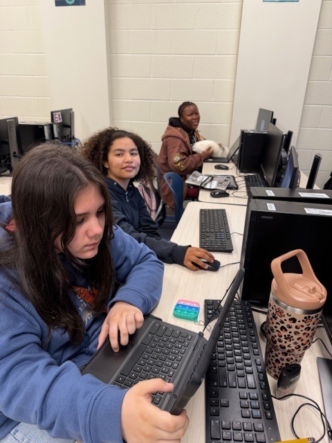 Three girls sit at their computers in a classroom. The girl closest to the camera looks at her Chromebook screen and the other two girls look at the camera and smile.