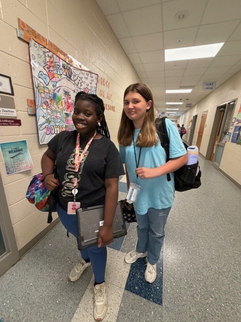 Two girls stand next to each other in the hallway and smile for the camera.