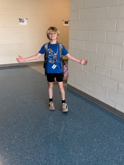 A boy stands in the hallway with his arms open smiling at the camera.