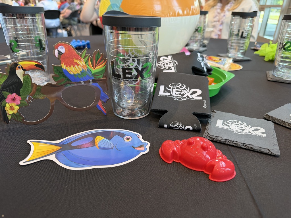 Items for the teachers are arranged at a table to include a Lex2 tervis tumbler, a Lex2 coozie, a Lex2 coaster, a cardboard fish, bird sunglasses, and a red crab sand toy.