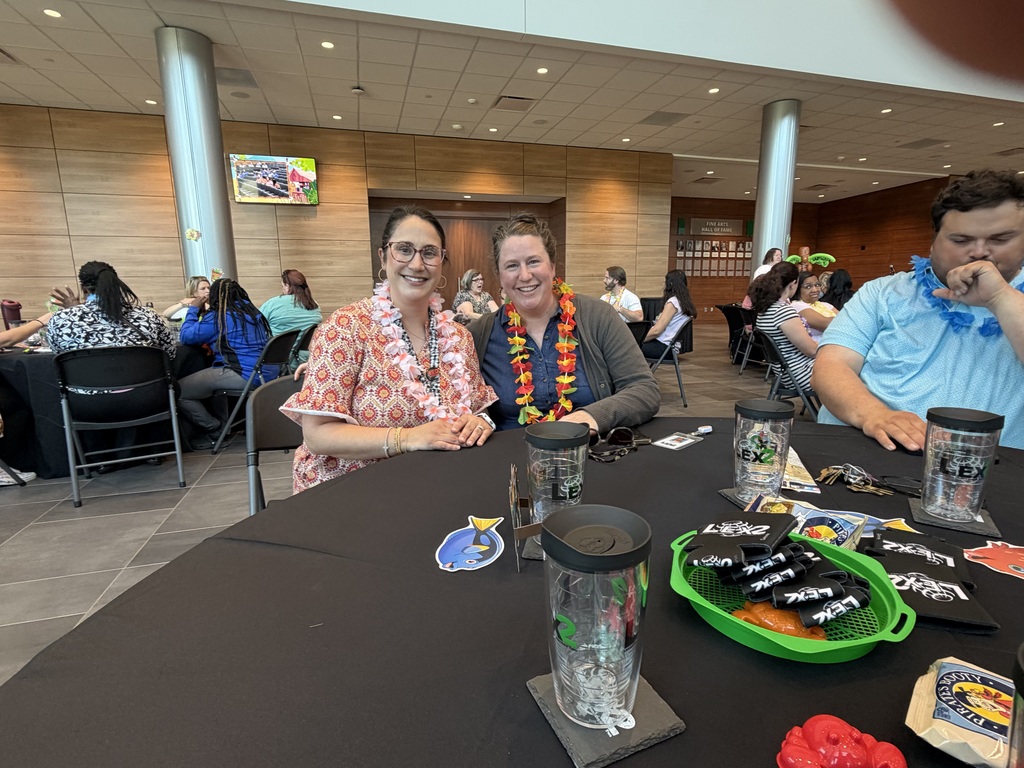 Two female teachers sit at a table and smile at the camera. Both teachers are wearing a floral lei.