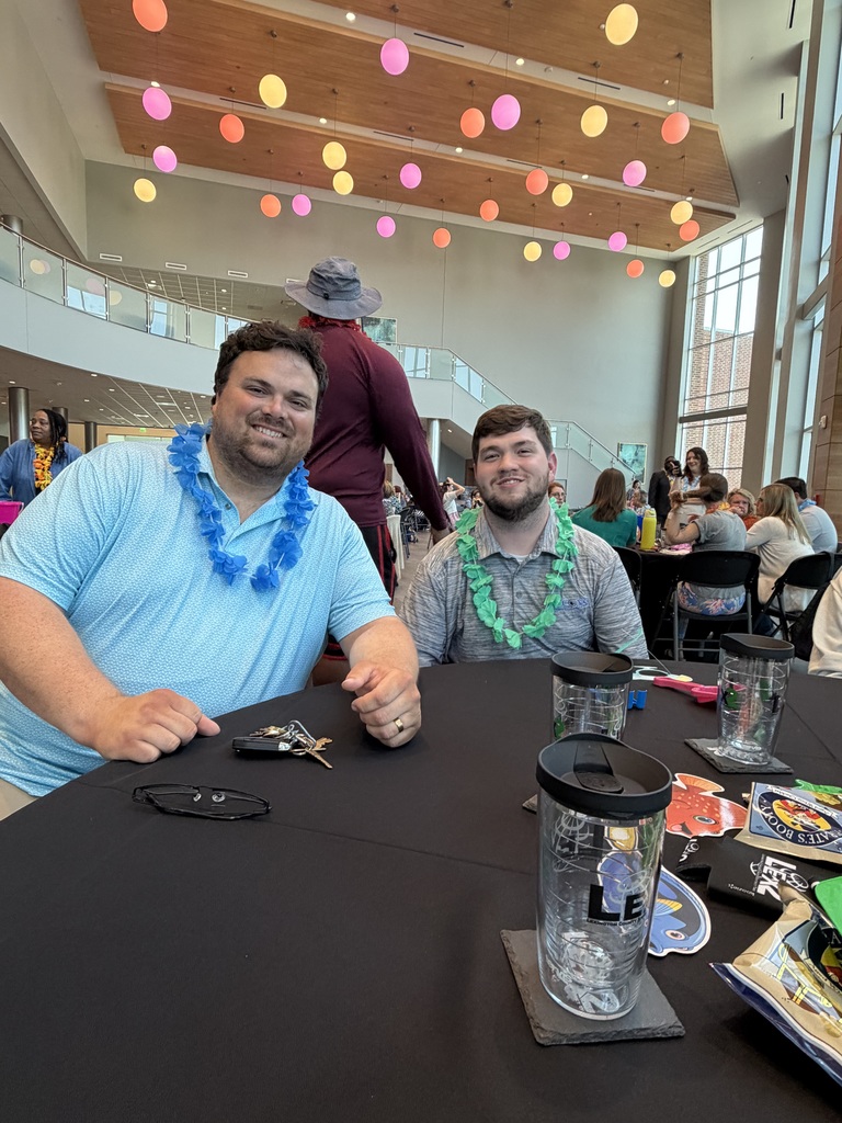 Two male teachers sit together at a table and smile at the camera.
