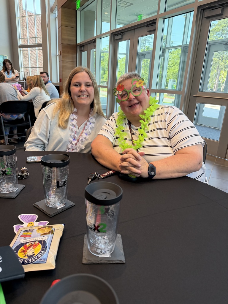 Two female teachers sit at a table and smile at the camera. Both teachers are wearing a floral lei.