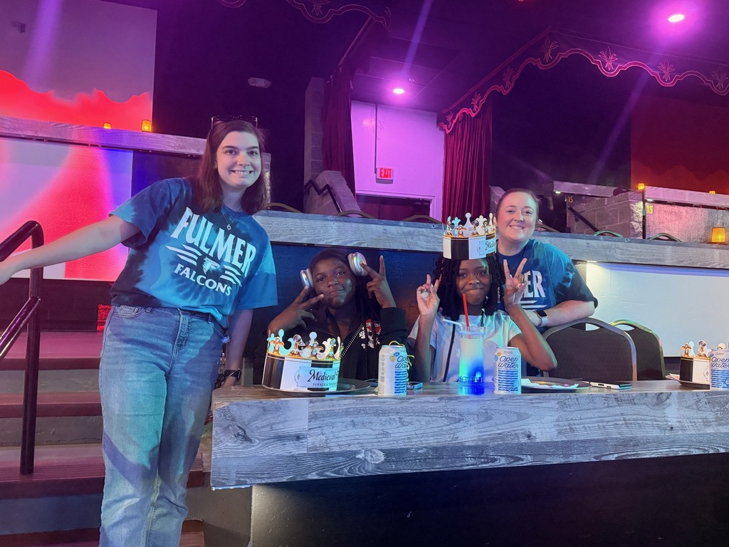 Students seated at tables waiting for the Medieval Times Show to begin. Two teachers are standing near the two students who are seated and holding peace signs.
