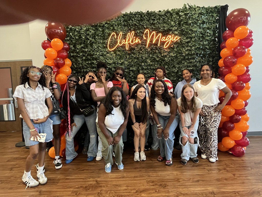 A group of female students and a few adults stand in front of a photo backdrop that has a lit up sign stating "Claflin Magic." Clafin University's school colors are in balloons that line from top to bottom of the backdrop.