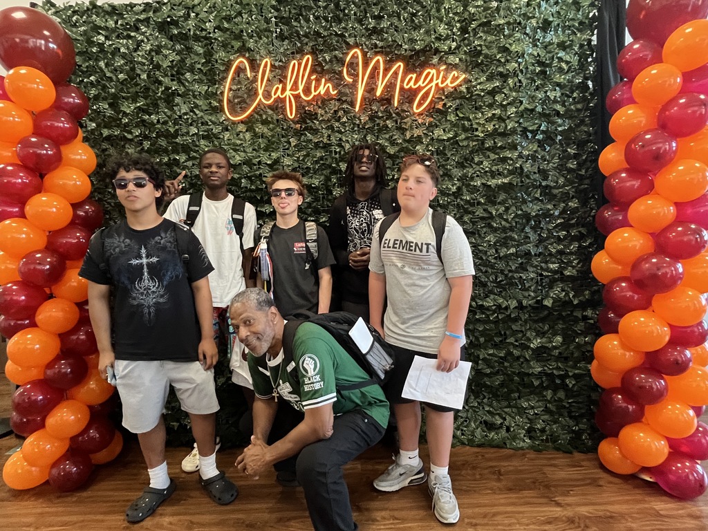 A group of female male and an adult stand in front of a photo backdrop that has a lit up sign stating "Claflin Magic." Clafin University's school colors are in balloons that line from top to bottom of the backdrop.