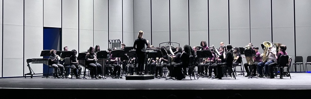 Students sit in chairs on a stage all dressed in black with instruments looking at the female conductor standing in the front middle.