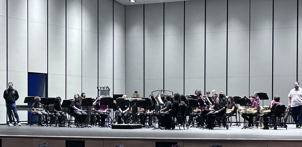 Students sit in chairs on a stage all dressed in black with instruments. The band instructors stand on both sides of the students. and are facing the crowd smiling.