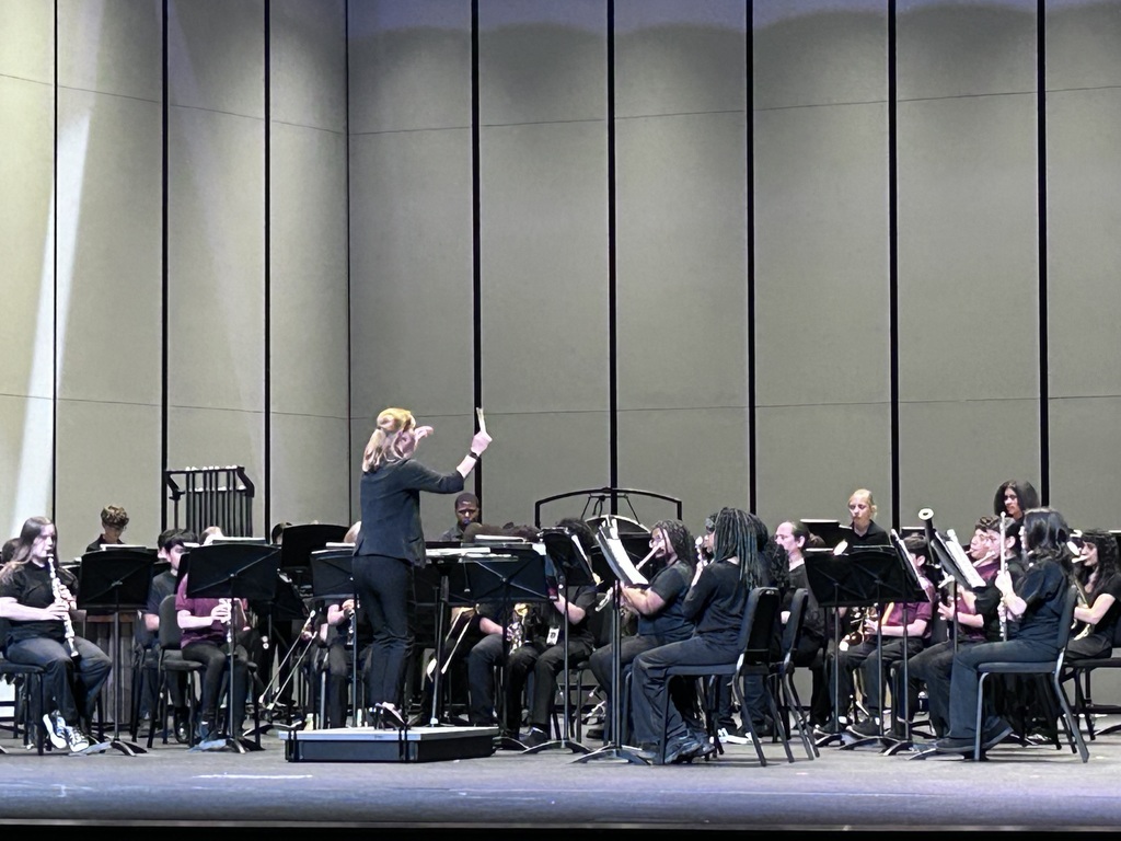 Students sit in chairs on a stage all dressed in black with instruments looking at the female conductor standing in the front middle.