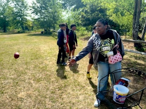 A female student stands outside and tosses a food item to an animal. Other students are standing in the background.