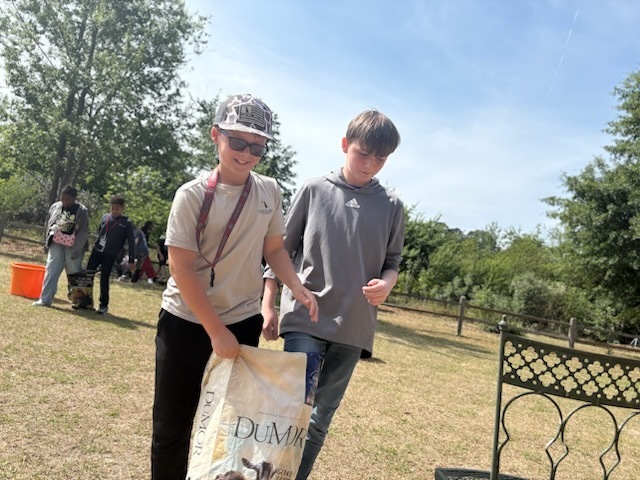 Two male students are outside walking side by side. One student is holding a bag of animal food for goats.