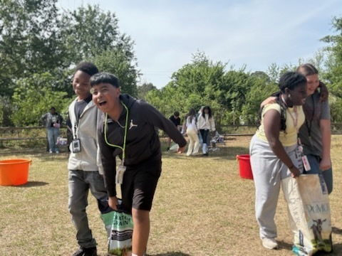 Two male students on the left of the image walk and laugh holding a bag of grain. Two female students on the right side stand next to each other and laugh and walk and one of them is holding a bag of food for goats.