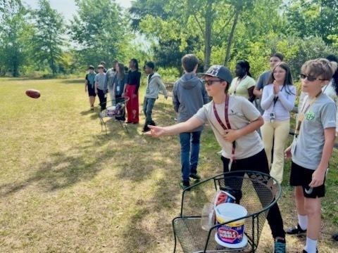 Students are standing outside in a line; one male student is shown tossing a food item to an animal.