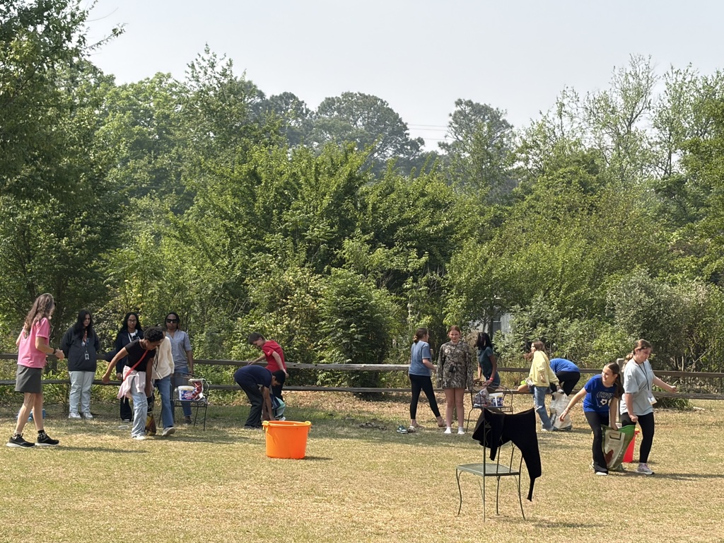 Several students are outside and are walking around with bags of animal food. An adult is standing near them overseeing what they are doing.