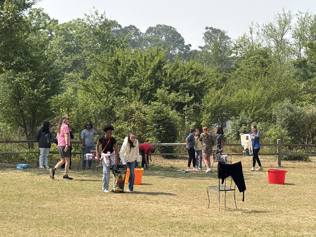 Several students are outside and are walking around with bags of animal food. An adult is standing near them overseeing what they are doing.