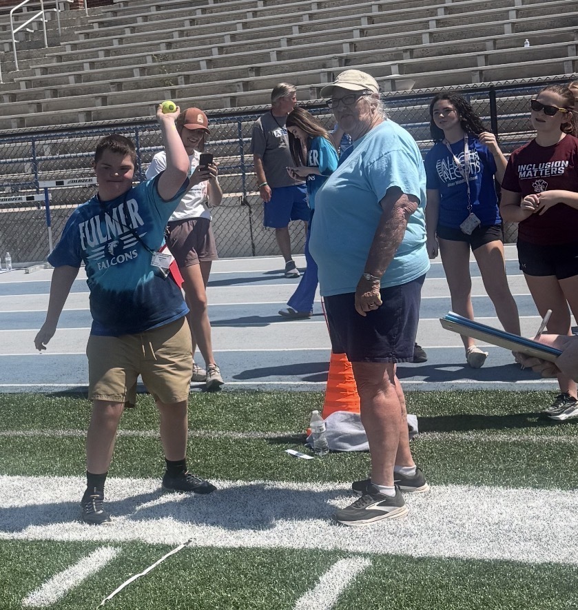 One student is standing on the sideline of the football field about to throw a tennis ball. Other adults and students are shown standing on the track in the background. One adult is next to the student throwing the ball looking at the place he will be throwing it.